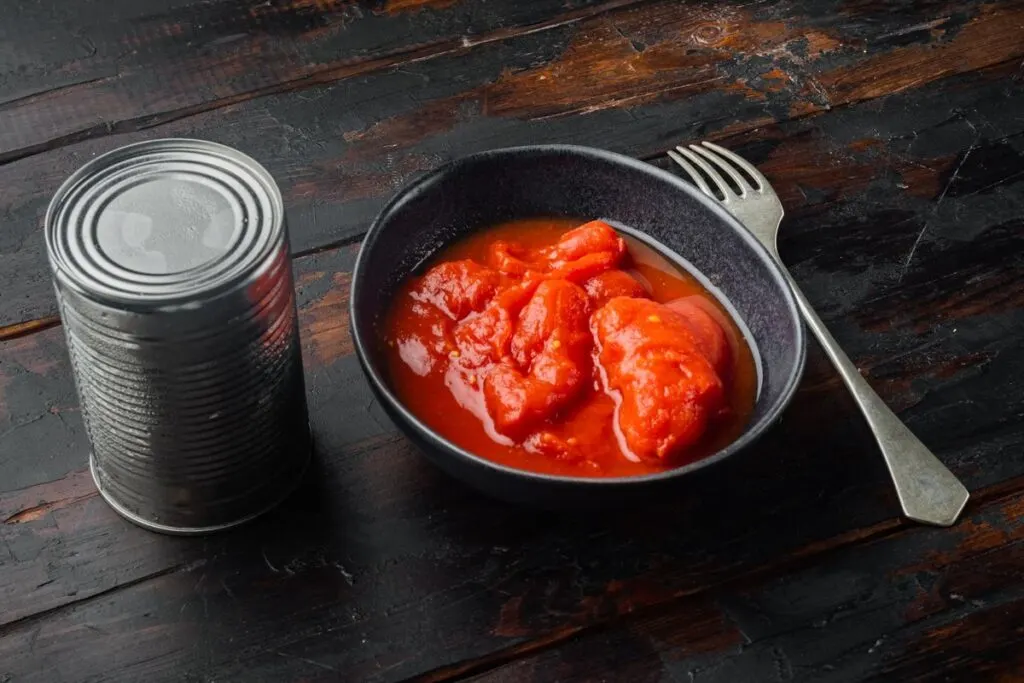An unopened can of tomatoes with a bowl of canned tomatoes in it.