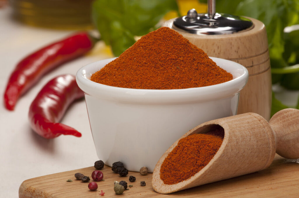 A bowl of chili powder with black pepper and chilis on the side.